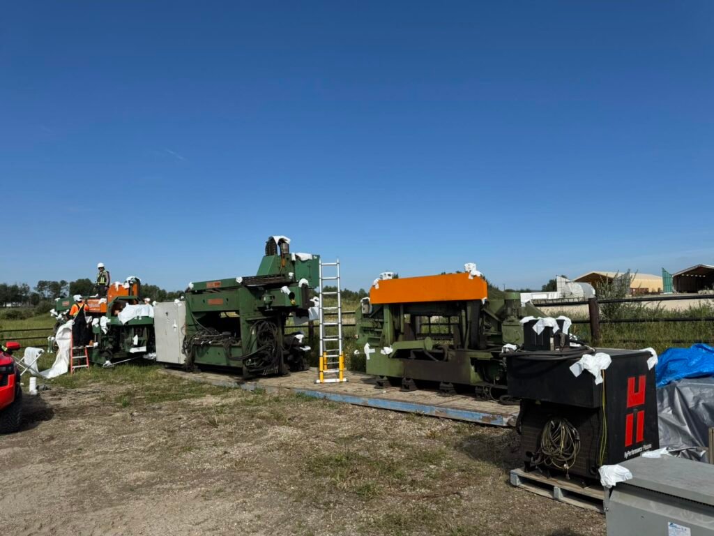 Workers apply white shrink wrap to green industrial machinery on a trailer.