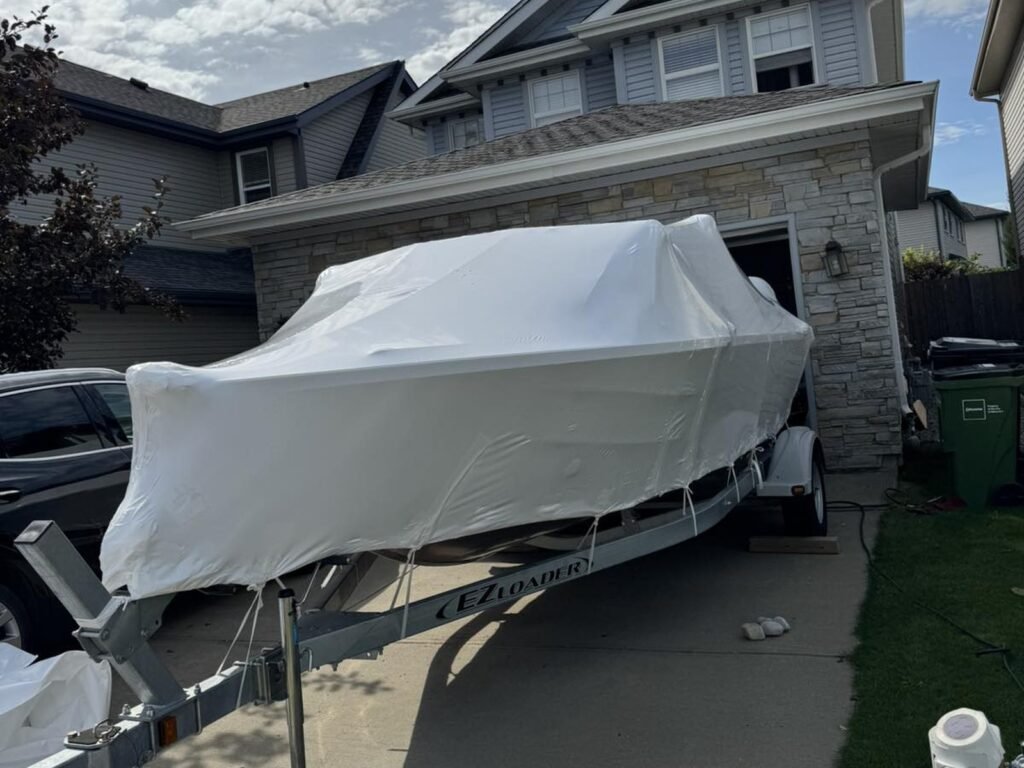 A black-and-white boat on a trailer is fully covered in white shrink wrap in a driveway.