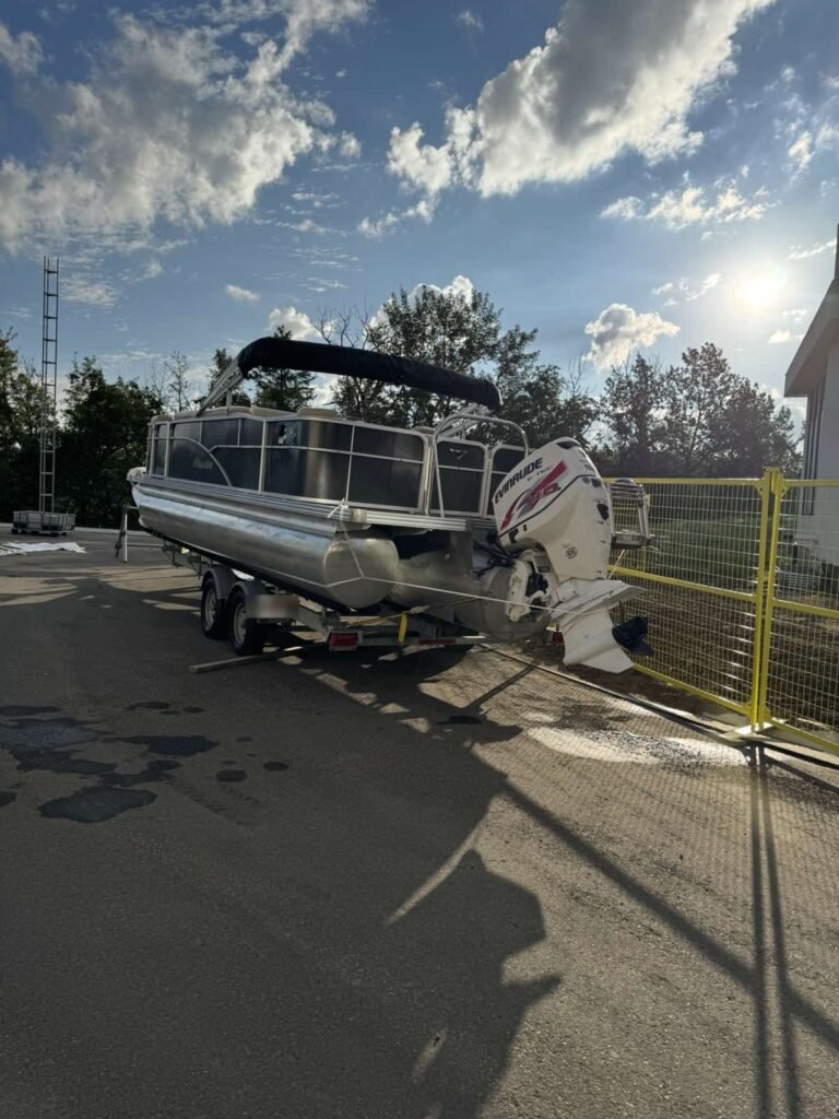 A pontoon boat on a trailer sits uncovered before shrink wrap.