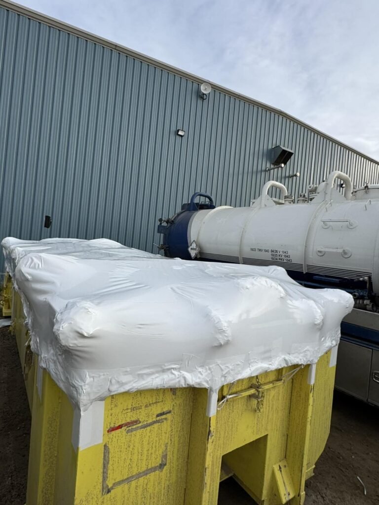 White shrink wrap seals the top of a yellow roll-off bin.